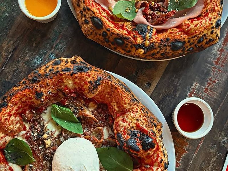 Two pizzas on a dark wooden table with a pot of honey and a dip, finished with basil and burrata.