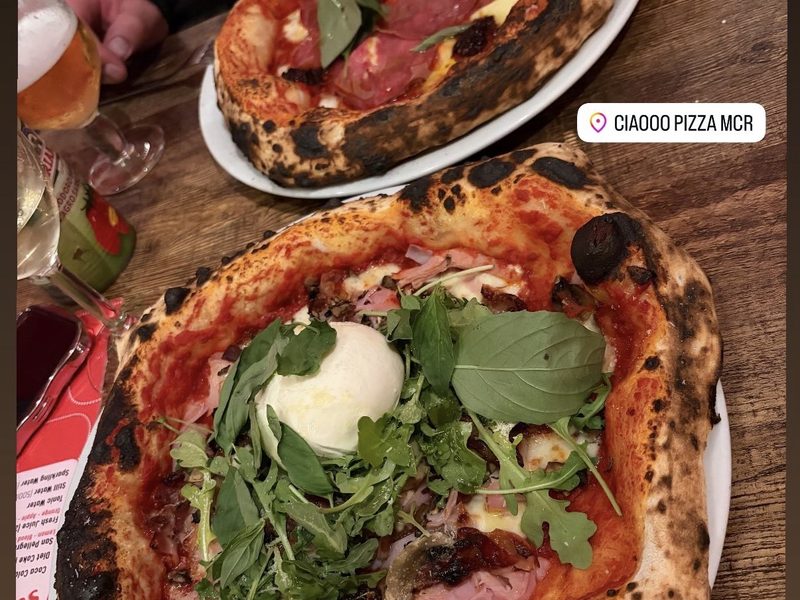 A pizza with rocket, cured ham and burrata beside a glass of beer on a Ciaooo table setting.